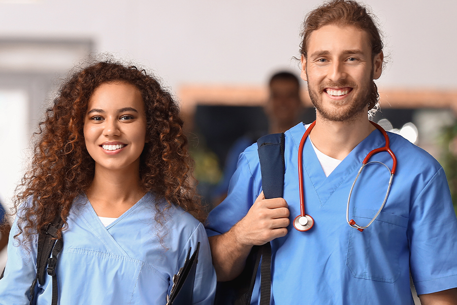 Two smiling students in blue scrubs and backpacks stand together, with one wearing a red stethoscope around their neck.