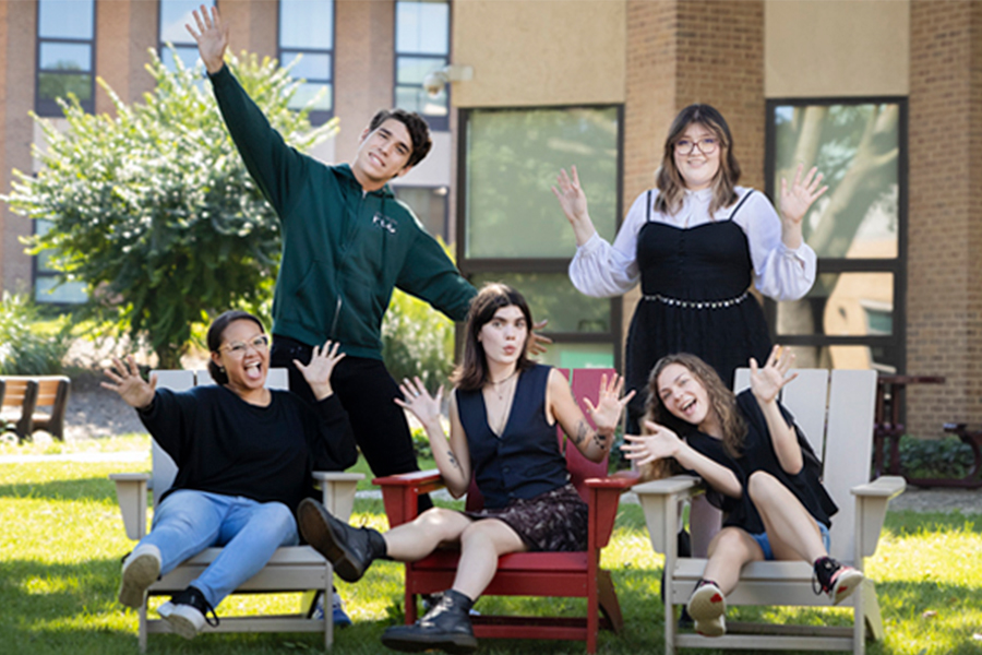 A group of five students posing playfully in Adirondack chairs on a sunny campus lawn.