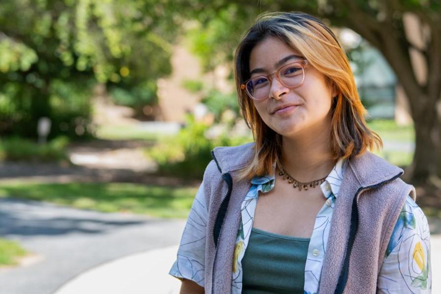 A young woman with glasses and highlighted hair smiling outdoors in a park-like setting.