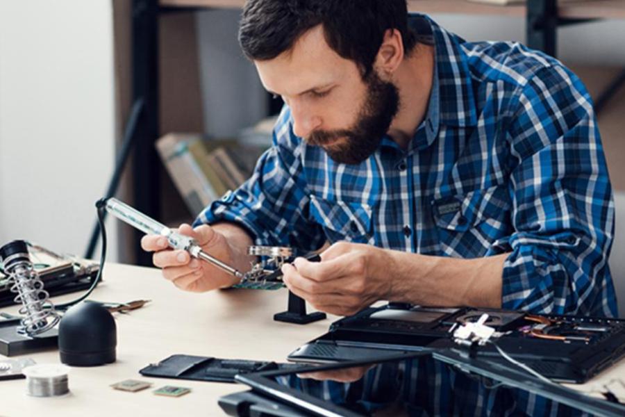 A man with a beard and a blue plaid shirt carefully solders a circuit board on a desk filled with electronics and tools.